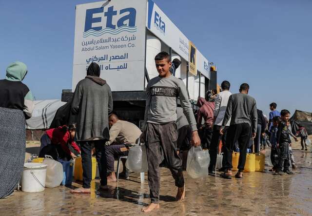 A man carrying two large plastic bottles of water from a water truck while others behind him stand in a line.