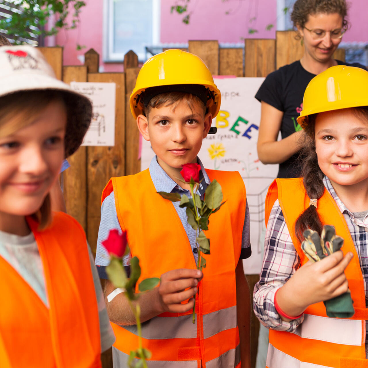 Ein Junge und zwei Mädchen halten Rosen in ihren Händen und präsentieren ihr Hochbeetprojekt. Alle drei tragen eine Warnweste und eine Kopfbedeckung.