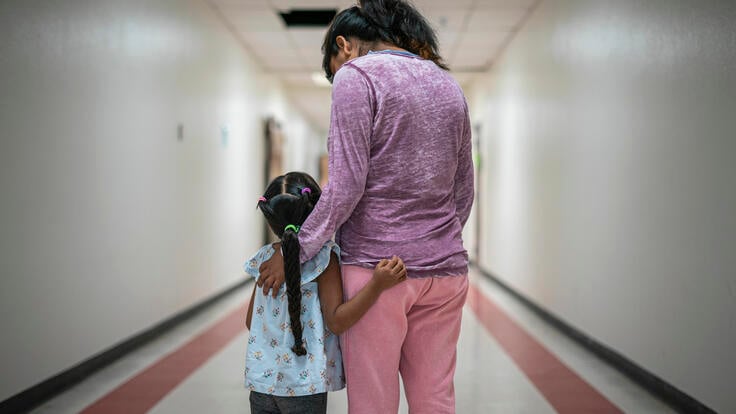 A Mexican mother wearing pink pants and a purple shirt stands with her arms around her young daughter, who has a long braid, in a hallway. They are asylum seekers in an IRC welcome center.