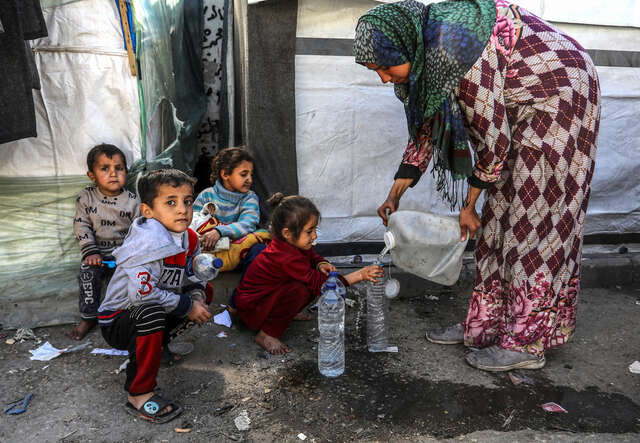 A woman in gaza fills water bottles as children sit on the street