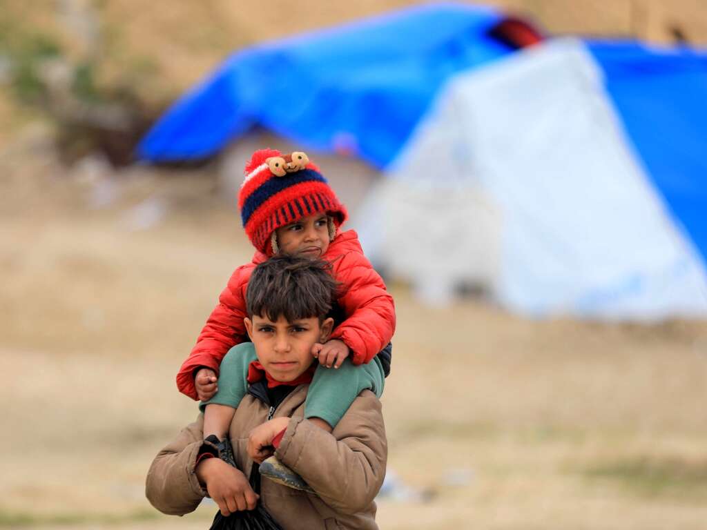 A young Palestinian boy carries his baby brother on his shoulders, outside of their makeshift shelter in Gaza.