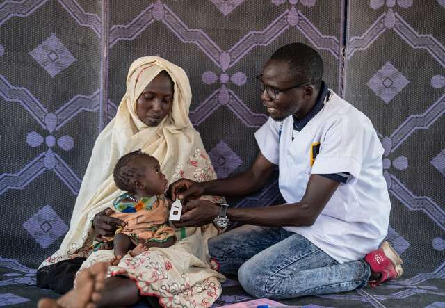 A young refugee is held by her mom as an IRC health worker screens her for signs of malnutrition.
