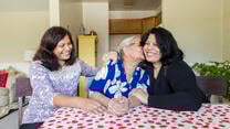 Three women, one older and two middle aged, sit at a table with a red and white table clothe. They are smiling and the older woman is kissing the woman on her left. They are all refugees.
